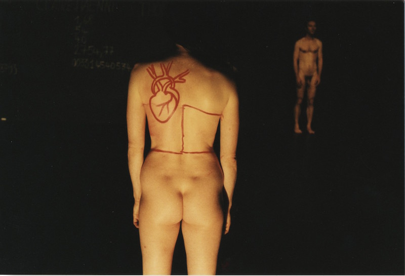 A woman's naked backside faces the audience. In red lipstick a drawing of a heart covers her shoulders.